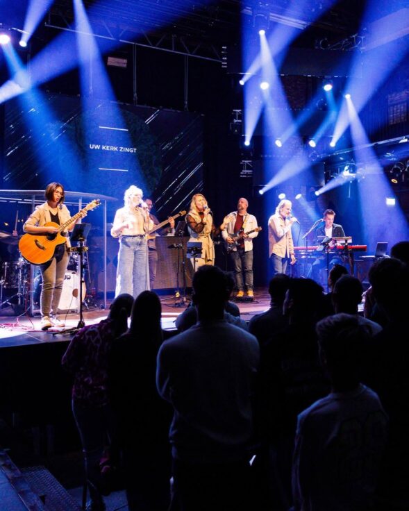 Worshipband van Mozaiek0548 speelt op het podium terwijl de zaal meezingt, met op het scherm de tekst Uw kerk zingt