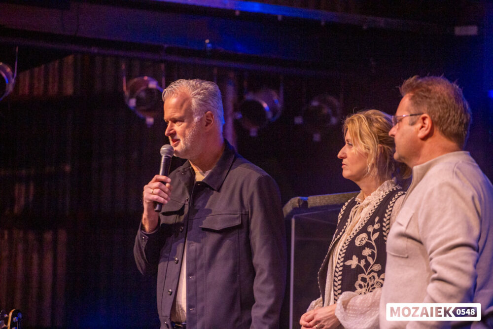Een man met microfoon staat met twee anderen op het podium tijdens een dienst van Mozaiek0548