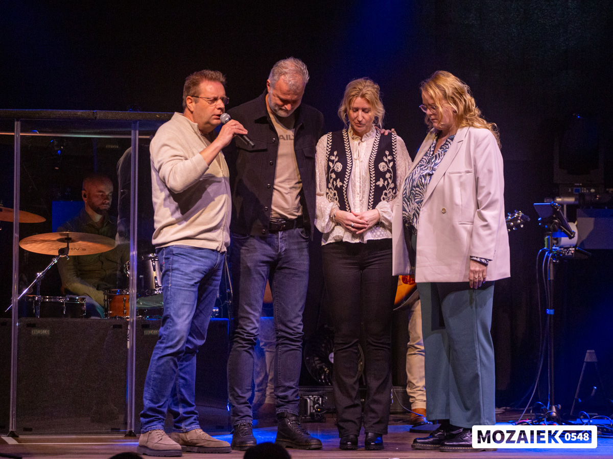 Vier mensen staan met gebogen hoofd samen te bidden op het podium tijdens een dienst van Mozaiek0548