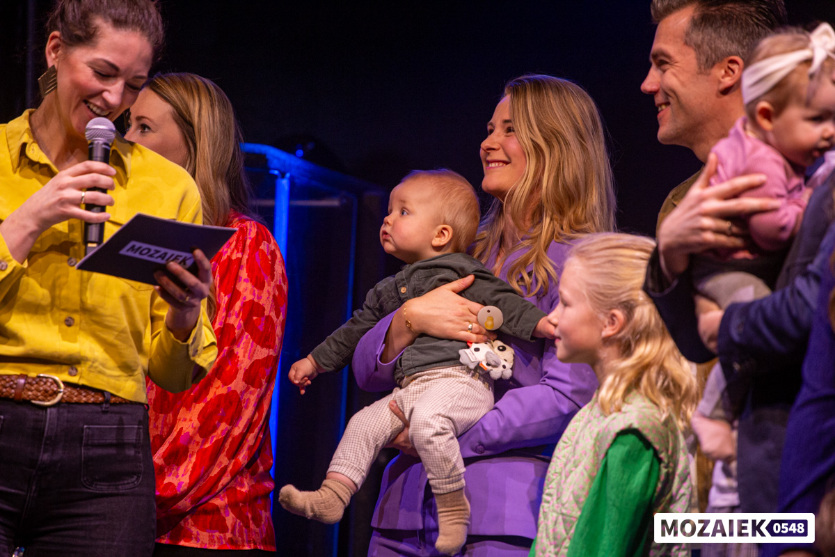 Een host met microfoon staat naast ouders die hun baby op de arm hebben tijdens een opdraagdienst