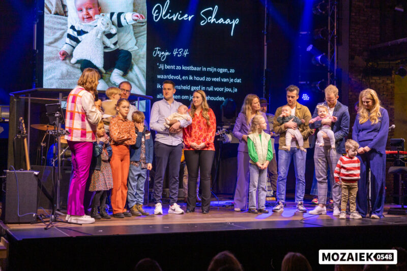 Gezinnen met hun kinderen op het podium met een bijbeltekst op het scherm tijdens een opdraagdienst