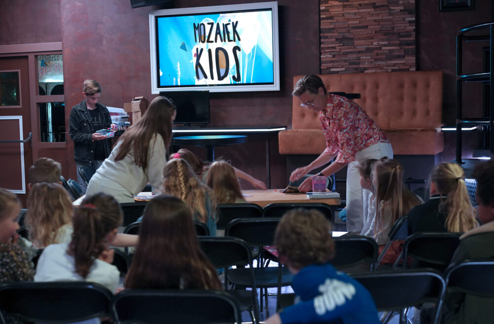 Kinderen zitten aan tafels in de ruimte van Mozaiek Kids terwijl het logo op het scherm staat