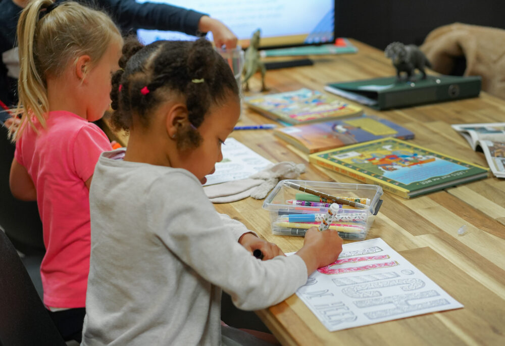 Twee meisjes werken met stiften aan een werkblad aan de tafel bij Mozaiek Kids