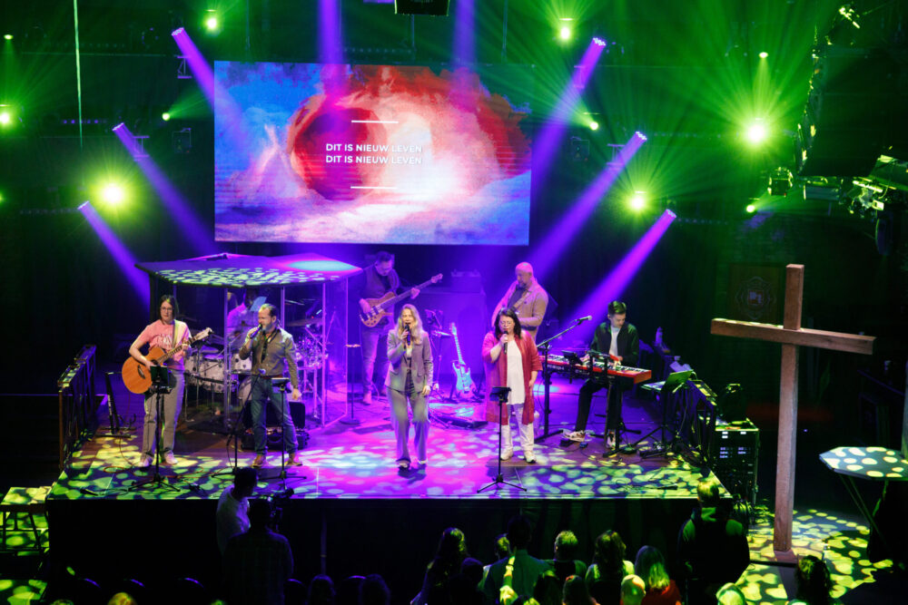 De worshipband op het brede podium onder groen en paars licht met het houten kruis ernaast