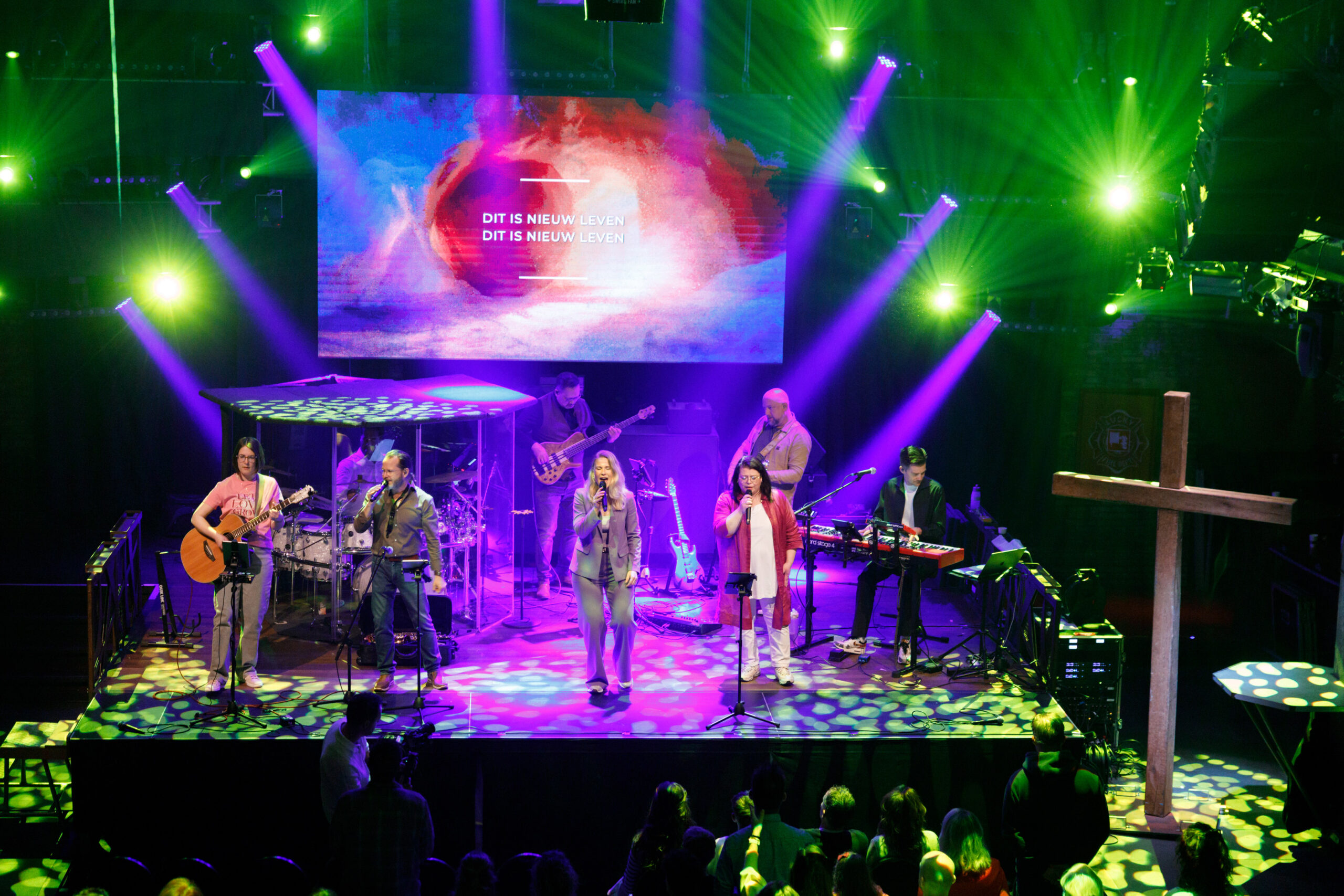 De worshipband op het brede podium onder groen en paars licht met het houten kruis ernaast