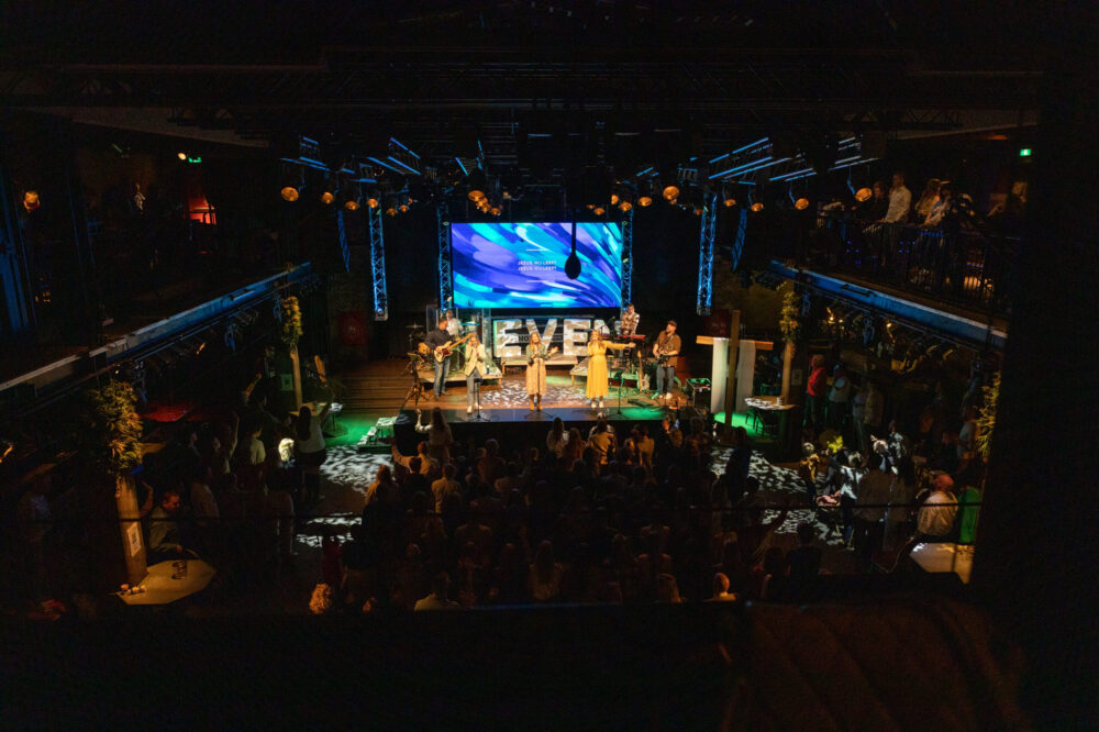 Overzicht van de volle zaal van Mozaiek0548 vanaf de achterzijde, met band op het podium en balkons vol bezoekers