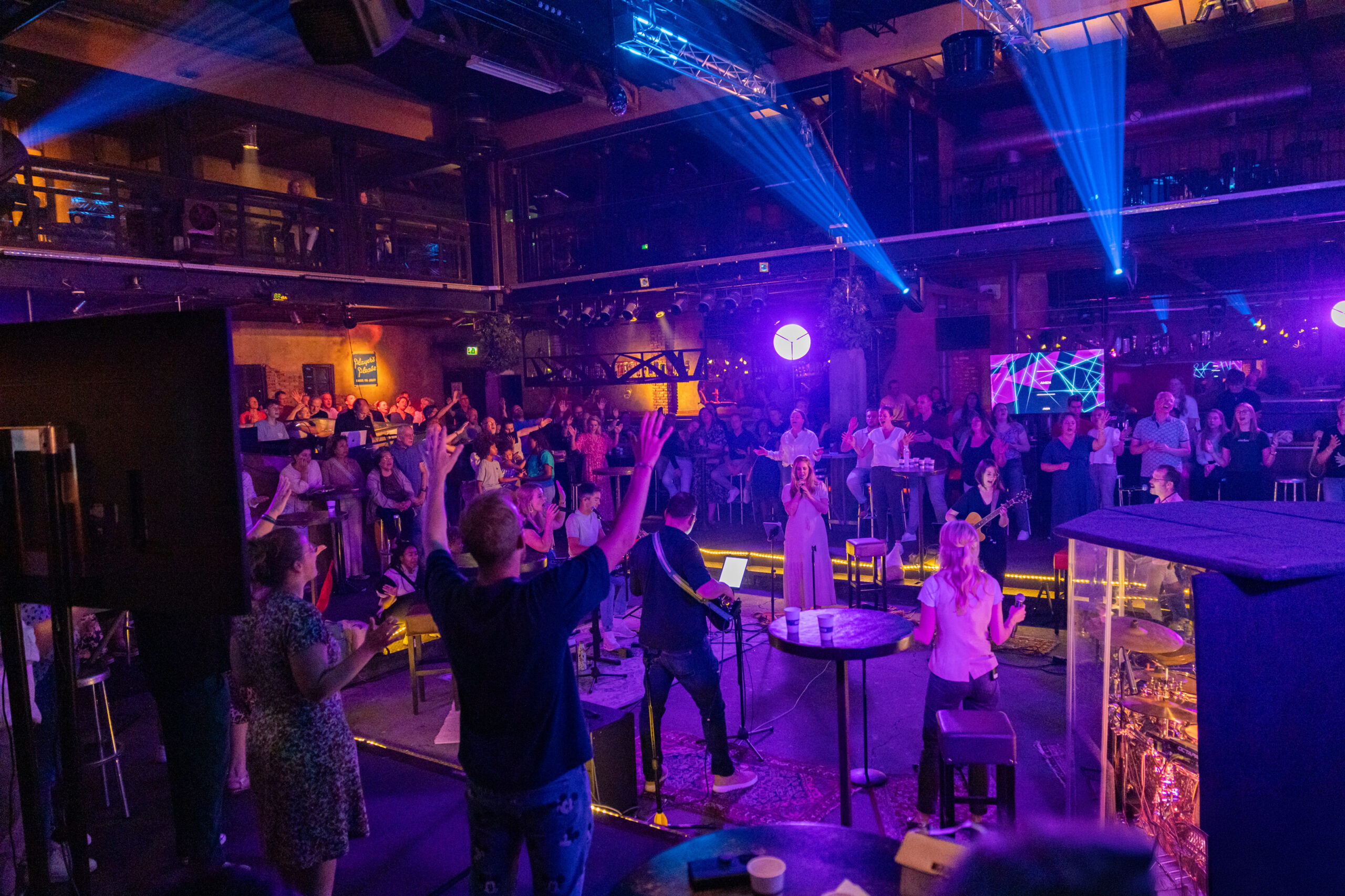 Bezoeker met opgeheven handen op het podium van Mozaiek0548, met de zaal en band eromheen