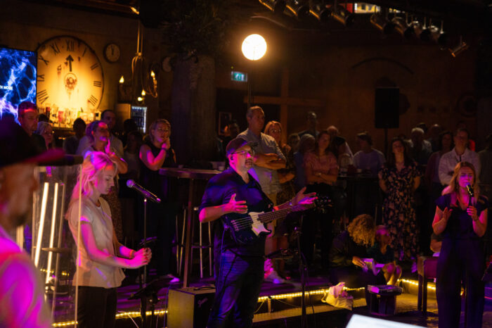 Bassist en zangeressen op het podium van Mozaiek0548 met bezoekers en de grote klok op de achtergrond