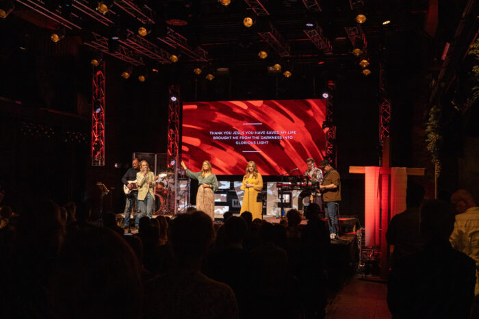 Overzicht van de zaal van Mozaiek0548 met worshipband op het podium en songtekst op het rode LED-scherm