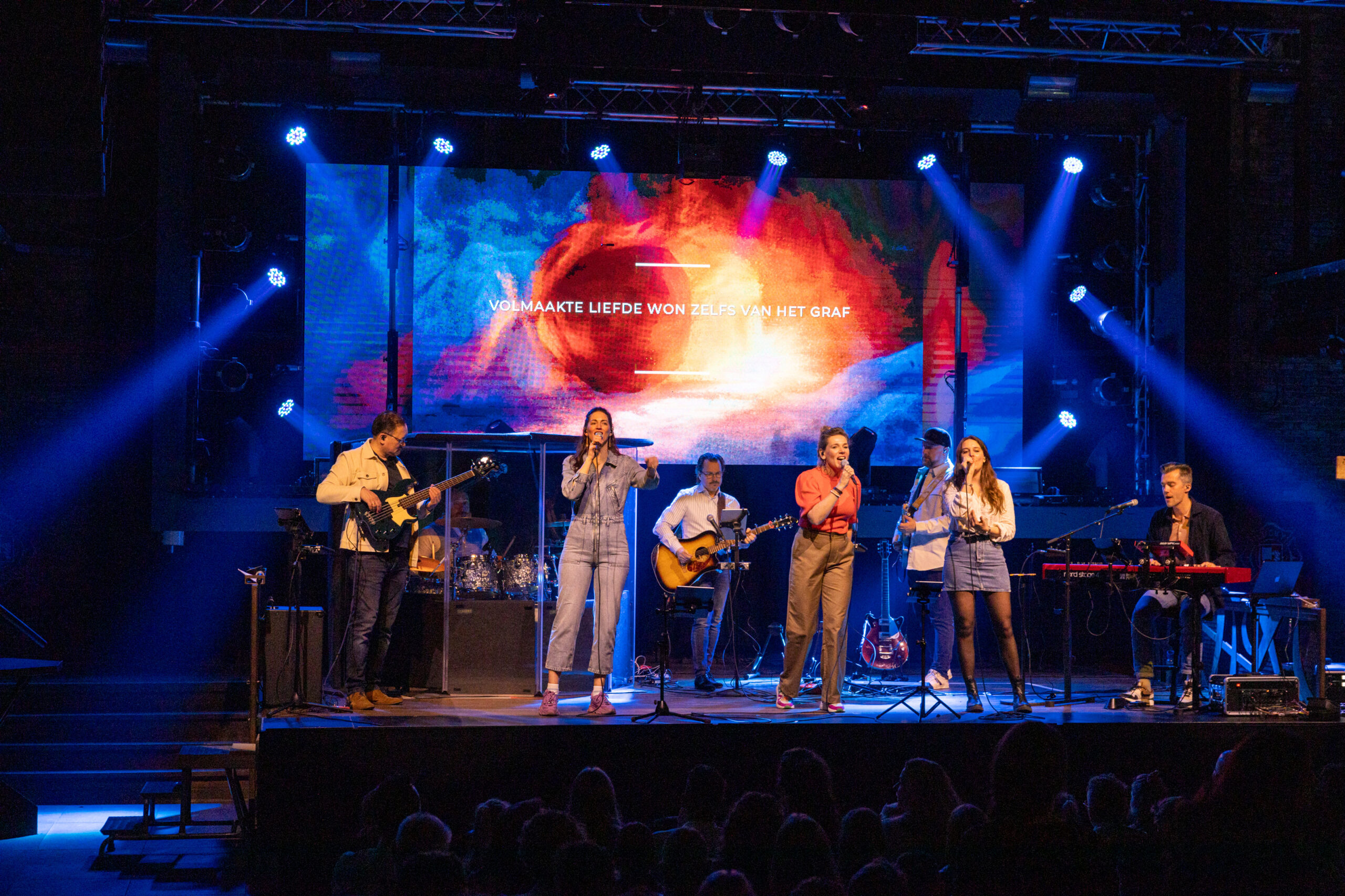 Worshipband van Mozaiek0548 op het podium met blauwe spots en Nederlandse songtekst op het LED-scherm